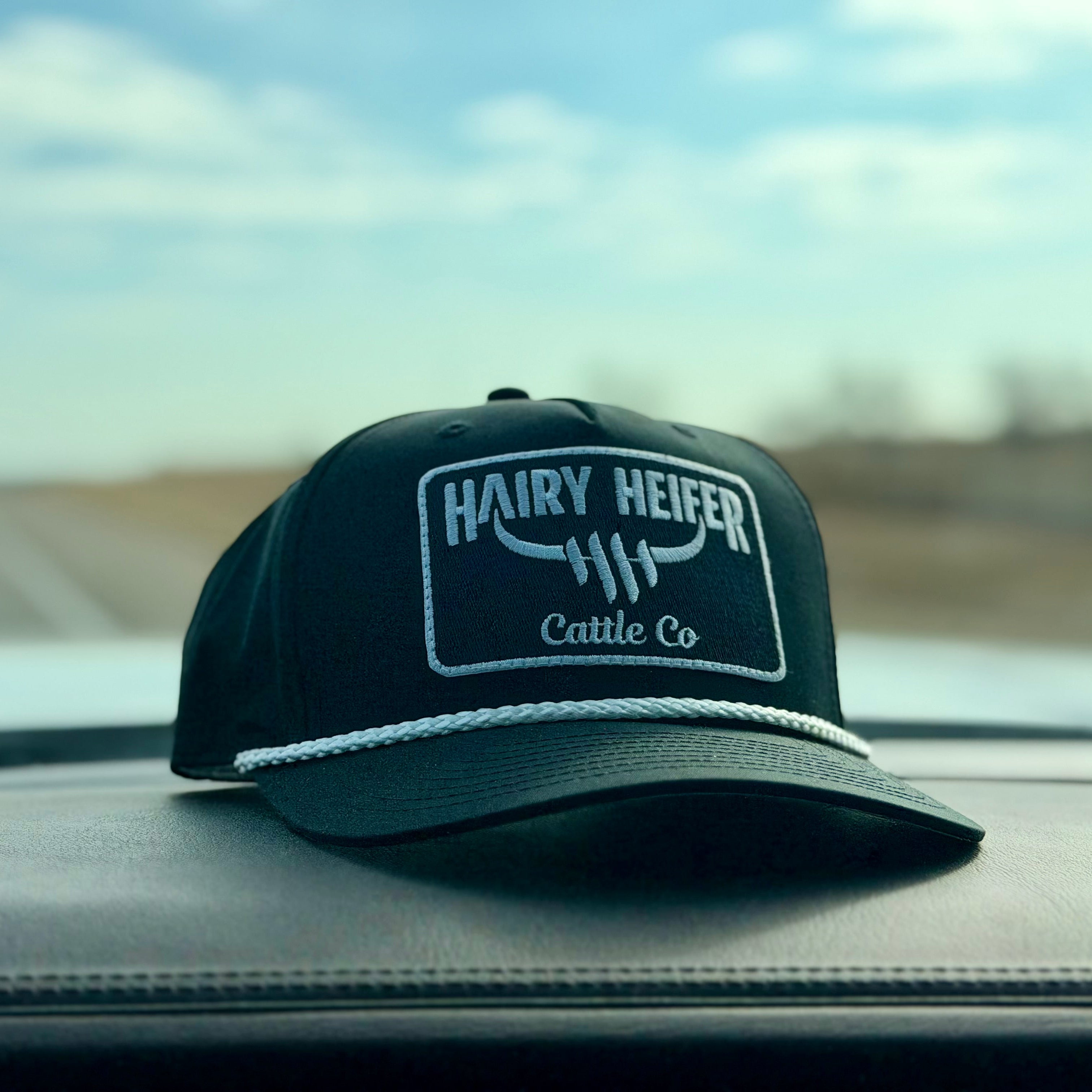Black cap with 'Hairy Heifer Cattle Co' logo on a car dashboard with a blurred road and sky in the background